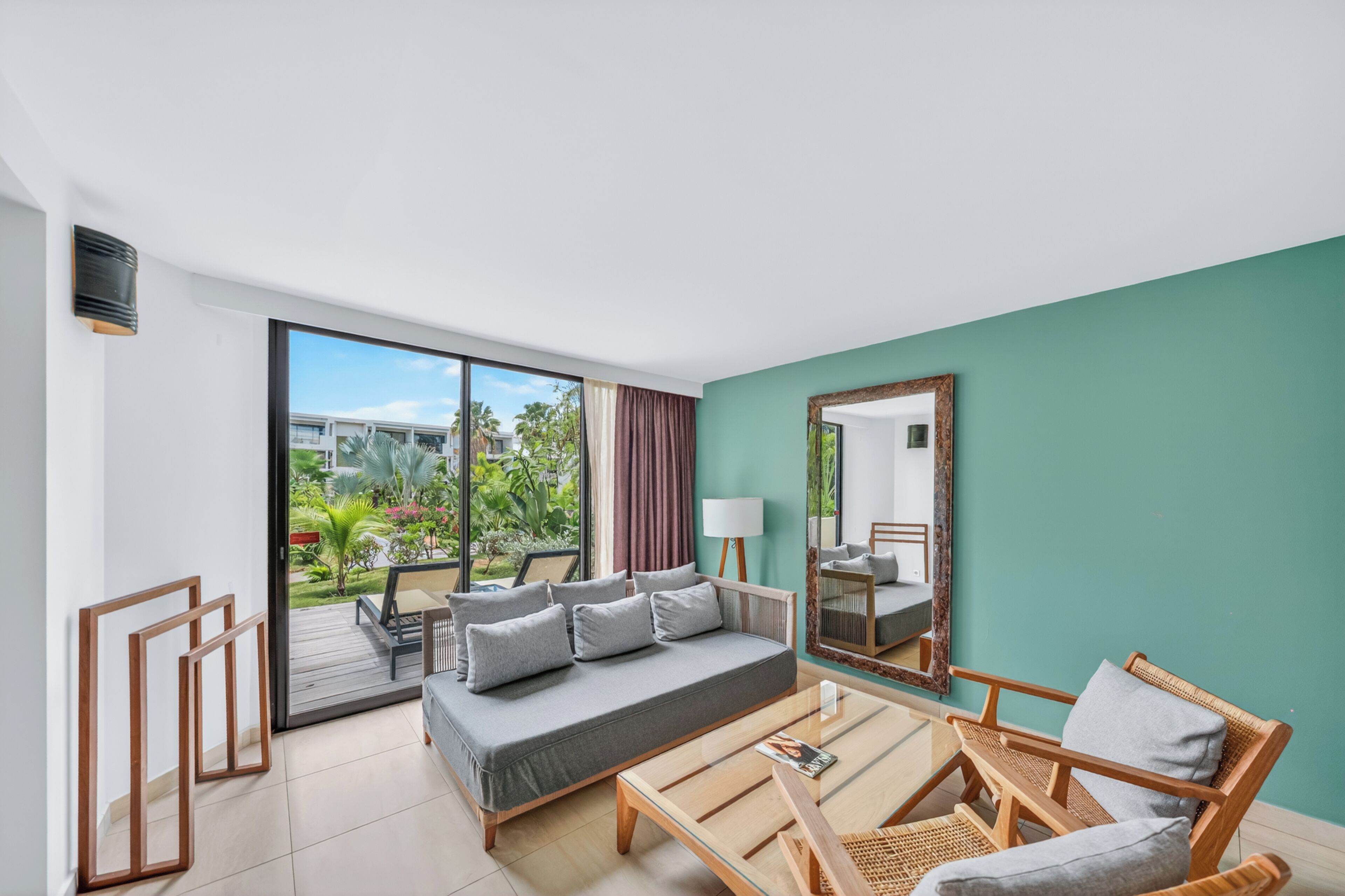 A modern hotel room with a large sliding glass door leading to a balcony, a sofa, a coffee table, and a mirror.
