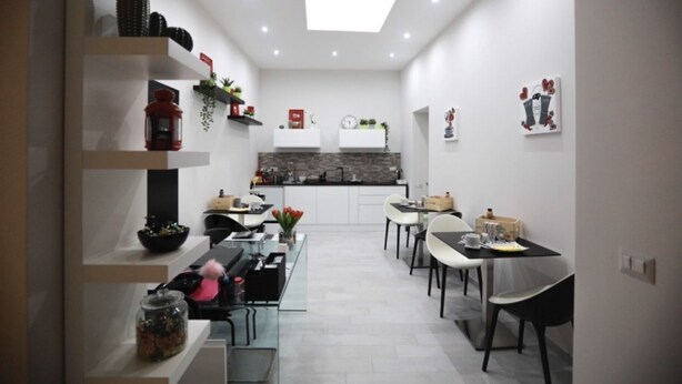 A modern kitchen with white cabinets, a black countertop, and a built-in stove. There is a dining area with white chairs and a table, and a small kitchen island with a sink and counter. - Chiaia Guest House (Naples)