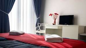 A neatly made bed with red and blue bedding, a white desk with a flat-screen TV, a chair, and a vase with flowers. - Chiaia Guest House (Naples)