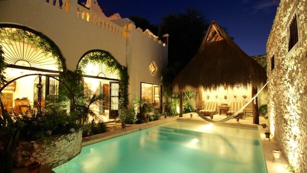 A pool area with a thatched-roof hut, a stone wall, and a building with arched windows and a sunburst light fixture. - Stay in Mérida's Grand Colonial Mansion w/ Private Pool (Centro)
