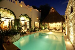 A pool area with a thatched-roof hut, a stone wall, and a building with arched windows and a sunburst light fixture.
