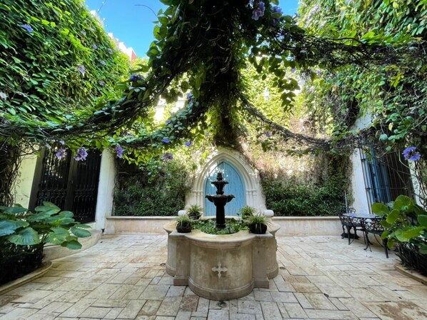 A courtyard with a fountain, surrounded by lush greenery and a stone floor.