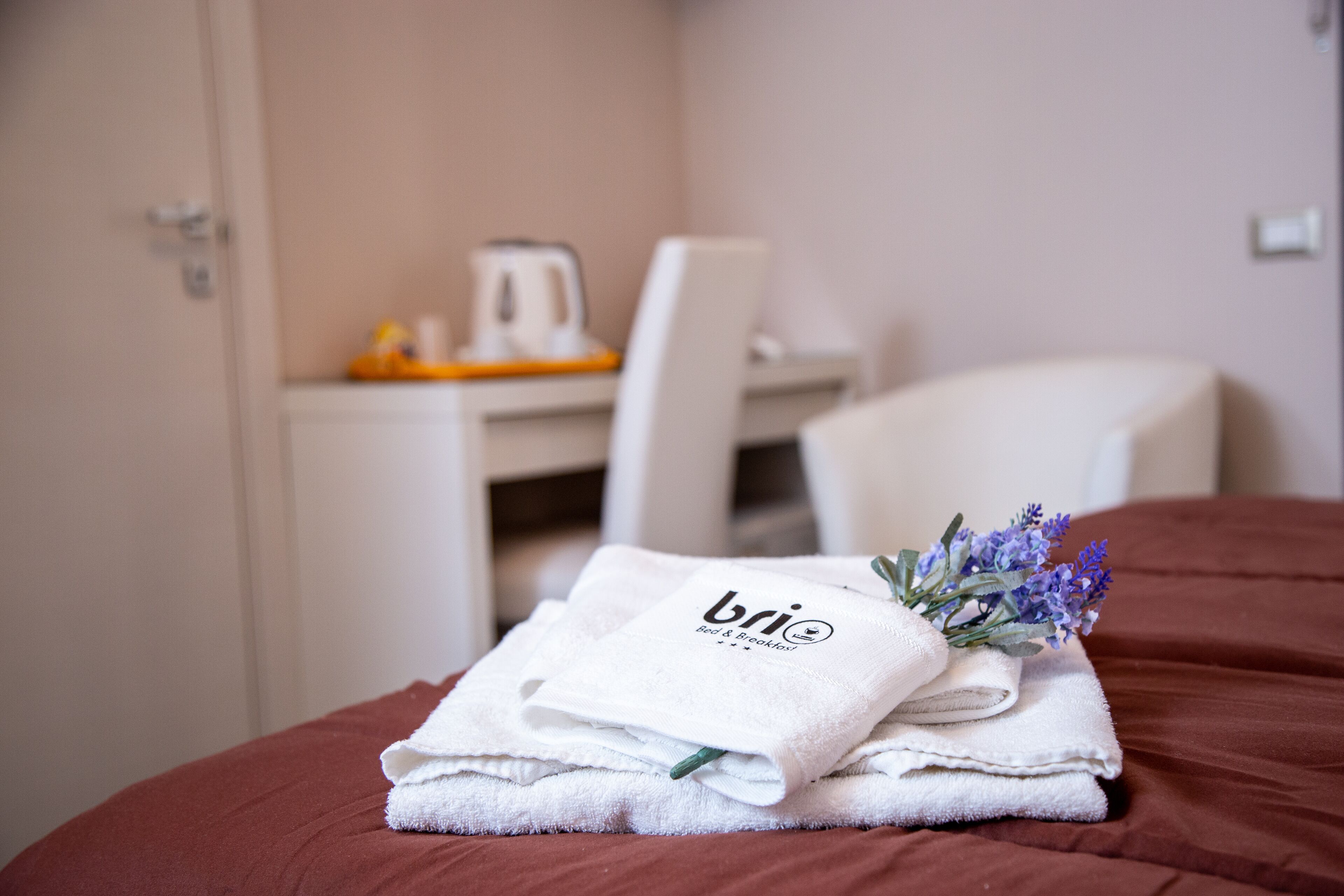 Un lit bien fait, recouvert d’une couverture marron, une pile de serviettes blanches ornées d’un logo et un bouquet de fleurs de lavande.