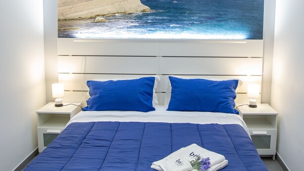 A bedroom with a large bed, blue bedding, two nightstands with lamps, and a painting of a coastal scene above the bed. - Brio Bed & Breakfast (Agrigento)