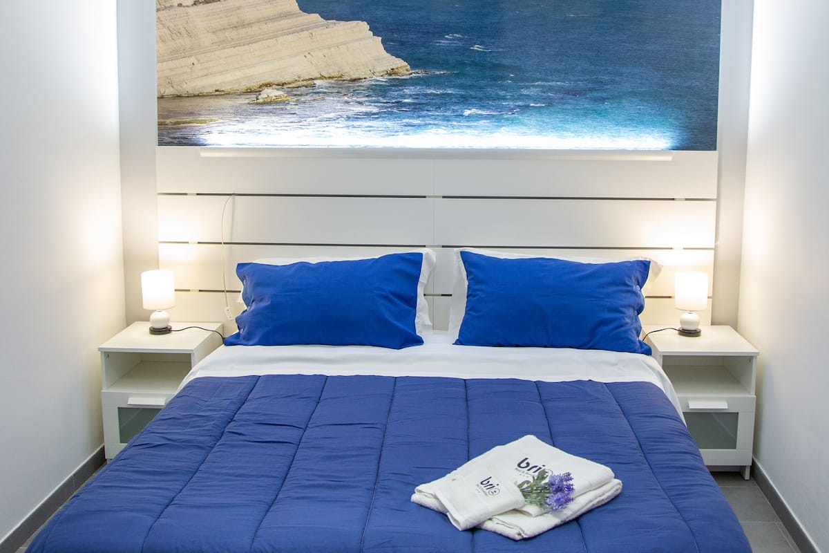 Un dormitorio con una cama grande, ropa de cama azul, dos mesitas de noche con lámparas y un cuadro con una escena costera sobre la cama.
