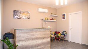A reception area with a wooden reception desk, a wall-mounted air conditioner, a framed photograph, and a small shelf with decorative items. - Brio Bed & Breakfast (Agrigento)