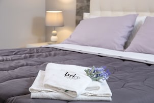 Una cama bien hecha con una toalla blanca doblada y un pequeño ramo de flores moradas sobre un edredón gris oscuro.