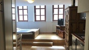 Een smalle gang met houten vloer, een bed, een radiator en een keuken met kastjes en een koelkast.