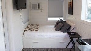 Een kleine, witte kamer met een bed, een aan de muur bevestigde televisie, een raam met zonwering en een klein tafeltje met een afstandsbediening.