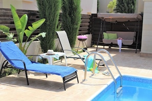 Un área junto a la piscina con una silla de descanso azul, una mesita con un sombrero y una silla colgante.