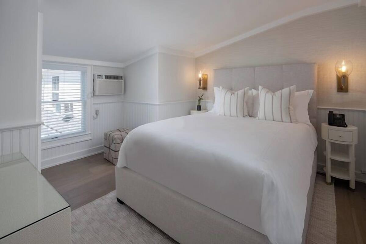 A spacious bedroom with a large bed, a bedside table, a nightstand, a window with blinds, and a wall-mounted air conditioning unit.