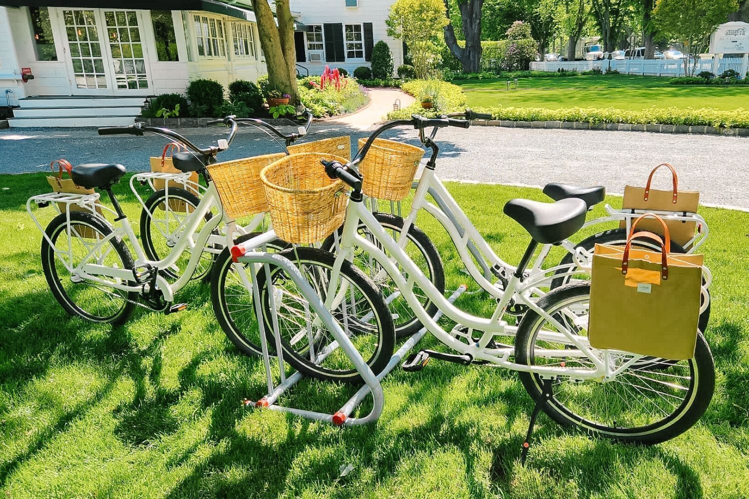 Três bicicletas estacionadas em um gramado com cestas e sacolas, em frente a uma casa com jardim bem cuidado e um caminho que leva à entrada.