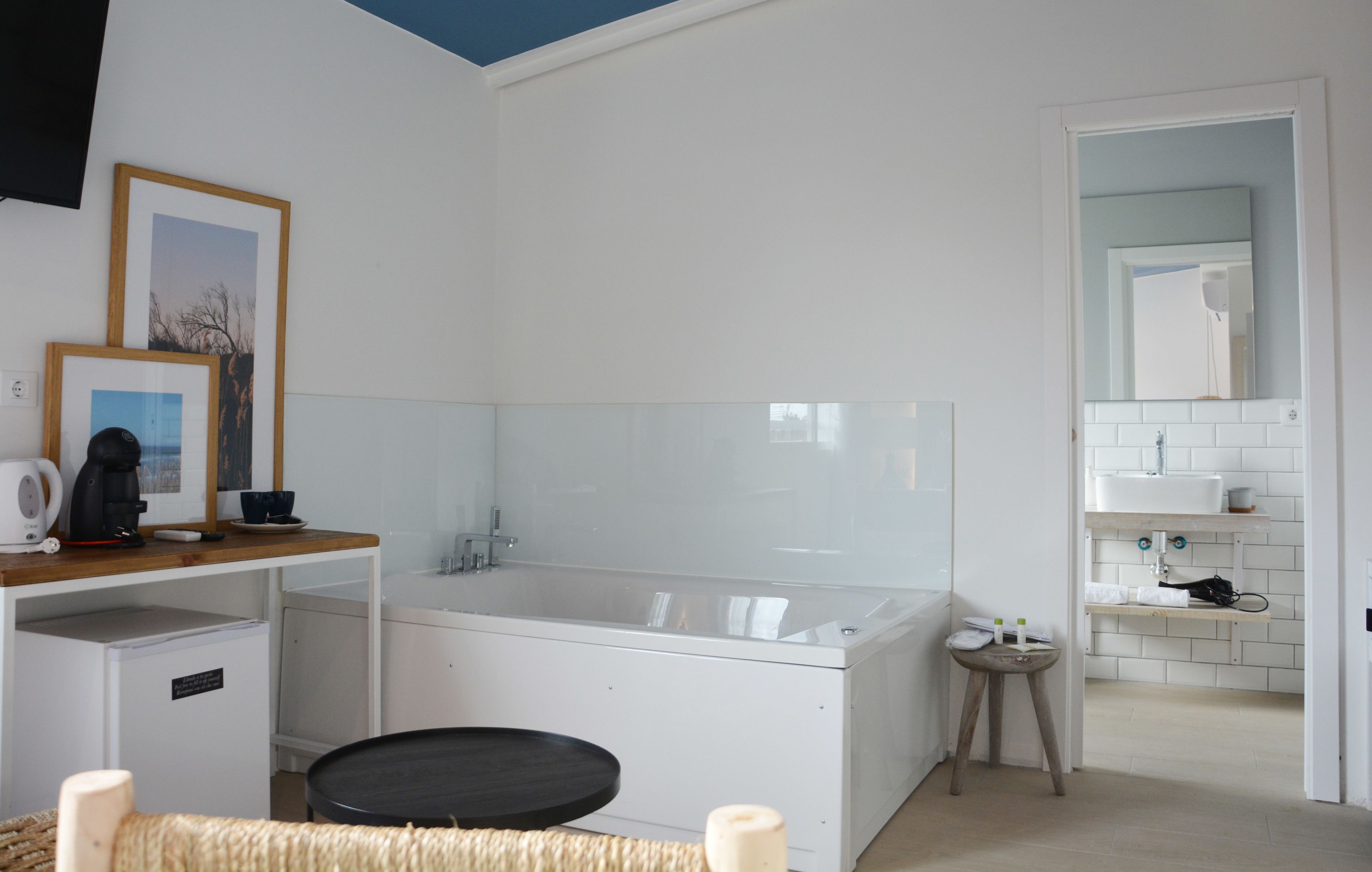 Ein modernes Badezimmer mit einer großen Badewanne, einem kleinen Waschbecken und einem Spiegel darüber. Eine Holzablage beherbergt eine Kaffeemaschine und Tassen, und ein kleiner Hocker bietet Platz für Pflegeprodukte.