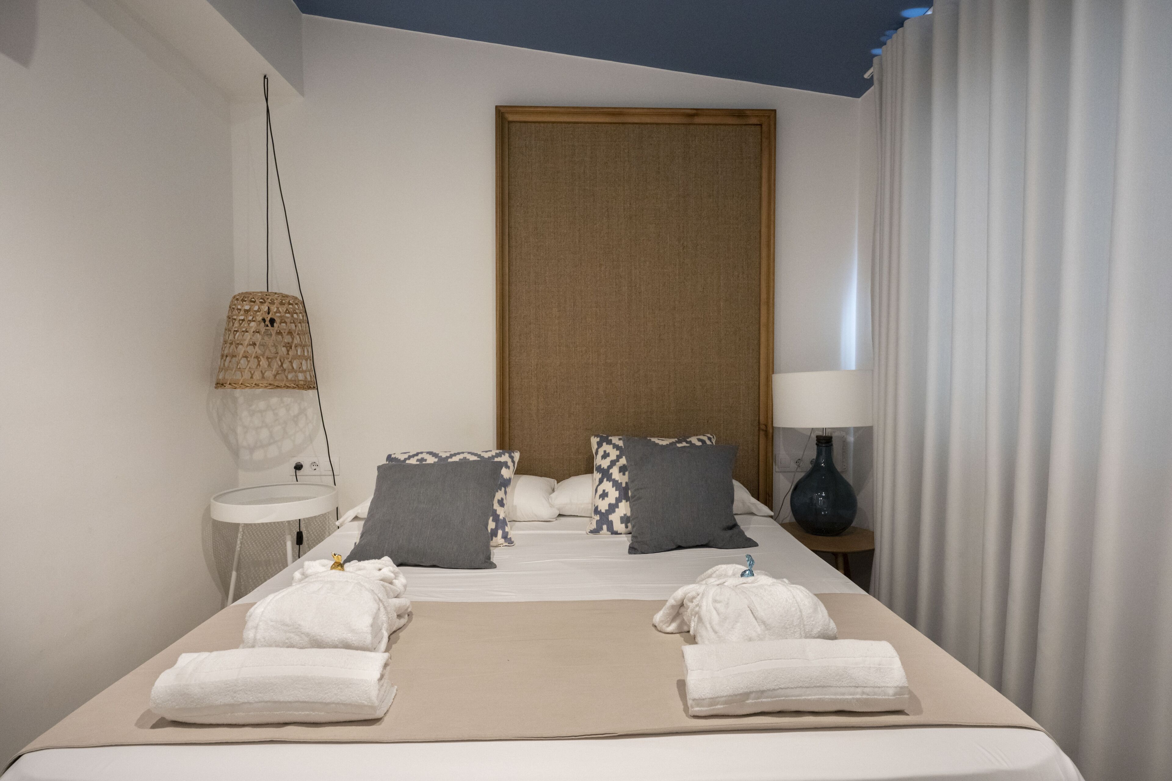 Una habitación de hotel con una cama, dos almohadas, una mesita de noche con una lámpara y una ventana con cortinas.