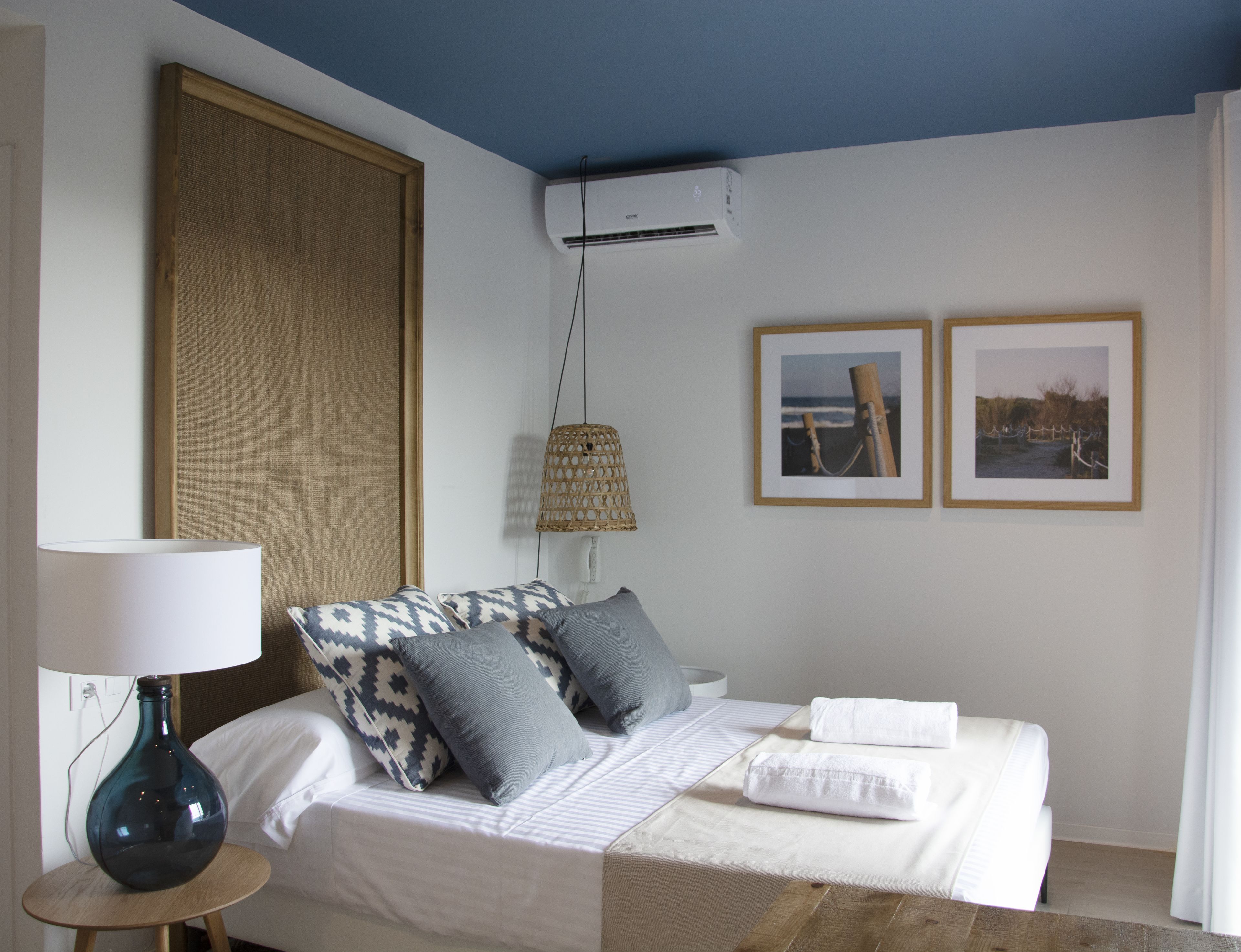 Habitación de hotel ordenada con una cama, una lámpara, aire acondicionado instalado en la pared y dos cuadros enmarcados.