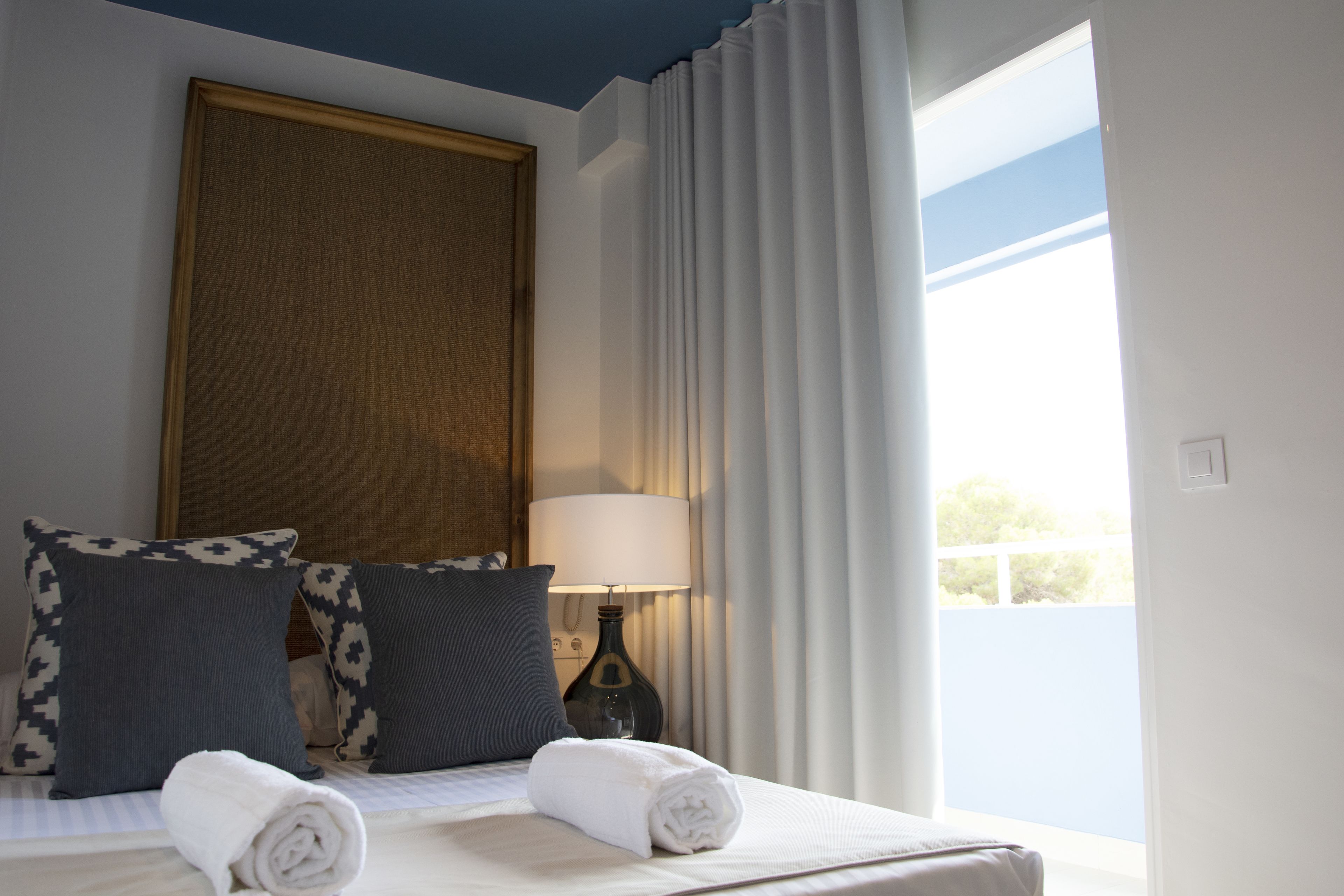 Una cama bien hecha con dos almohadas, una toalla blanca enrollada y una lámpara de noche en una habitación con un ventanal y cortinas.