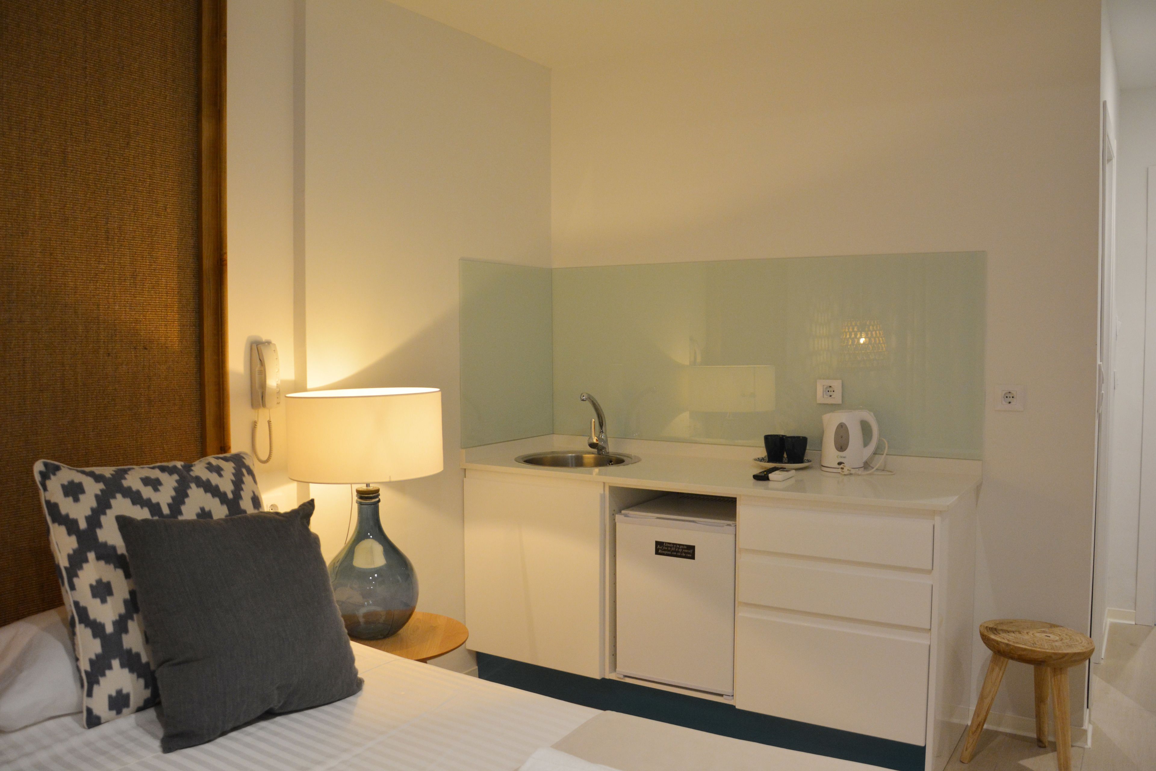 Ein Hotelzimmer mit Bett, Nachttisch mit Lampe, einer kleinen Küchenzeile mit Spüle, einem Spiegel und einem Hocker.