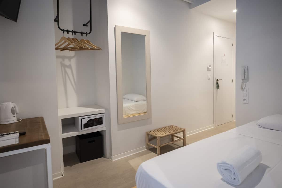 Une chambre d’hôtel comprenant un lit, une table de chevet avec une bouilloire, un miroir, une armoire et un petit tabouret.