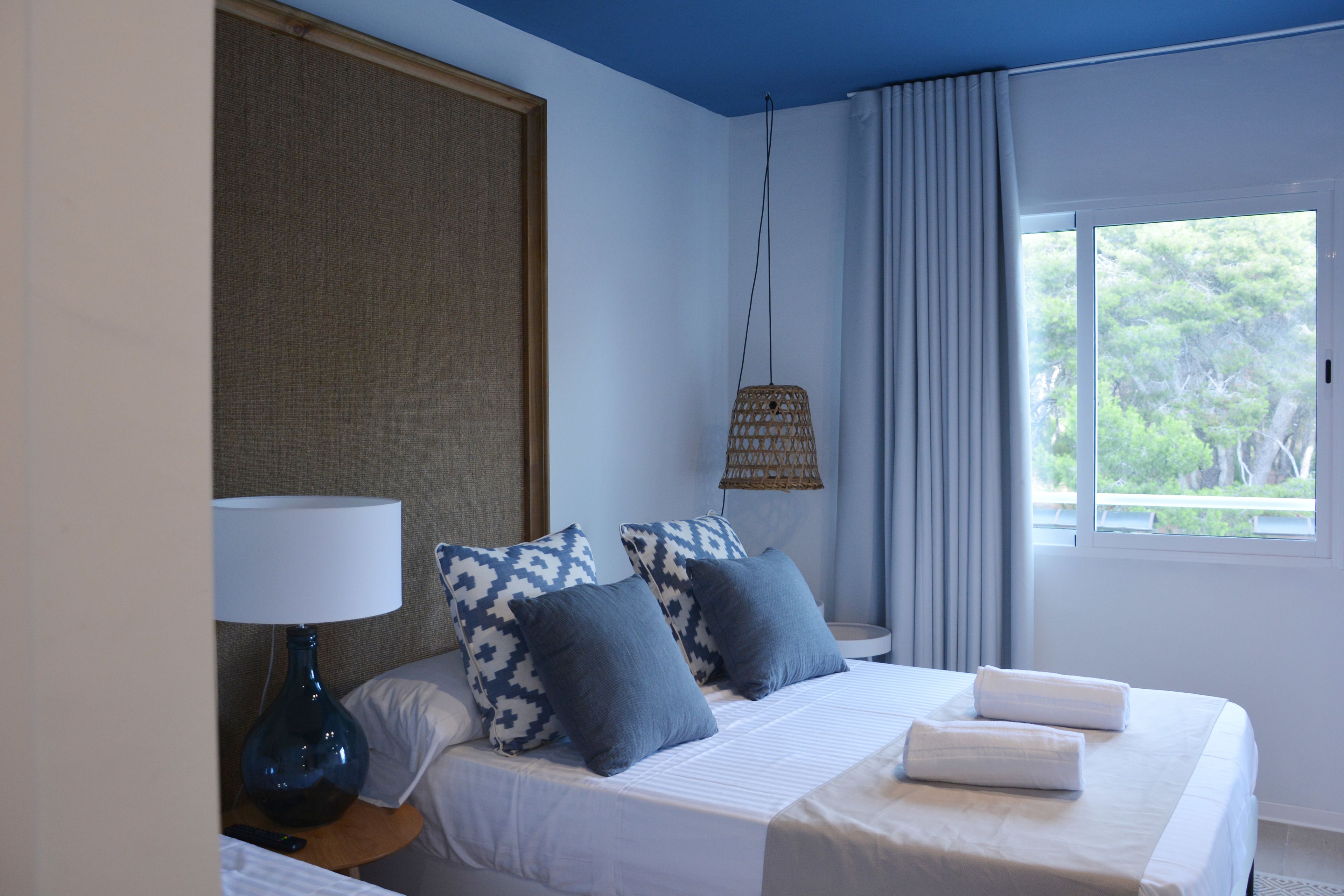 A hotel room with a bed, a lamp, a hanging lamp, a window with curtains, and a view of trees outside.