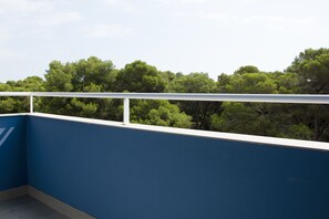 Un balcon avec un mur bleu et une balustrade blanche, donnant sur une zone boisée dense.