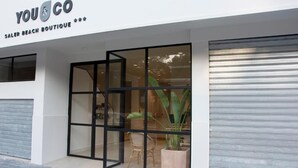 Eine Schaufensterfront mit Glastür und -fenstern, einem Schild mit der Aufschrift "YOU & CO SALER BEACH BOUTIQUE" und einer Pflanze im Innenbereich.