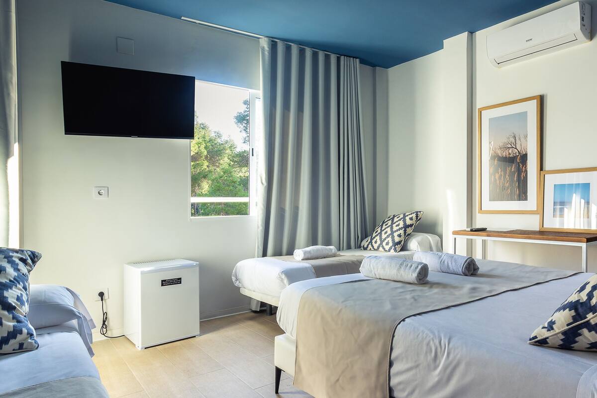 Une chambre d’hôtel avec deux lits, un téléviseur fixé au mur, un petit climatiseur et une fenêtre avec des rideaux.