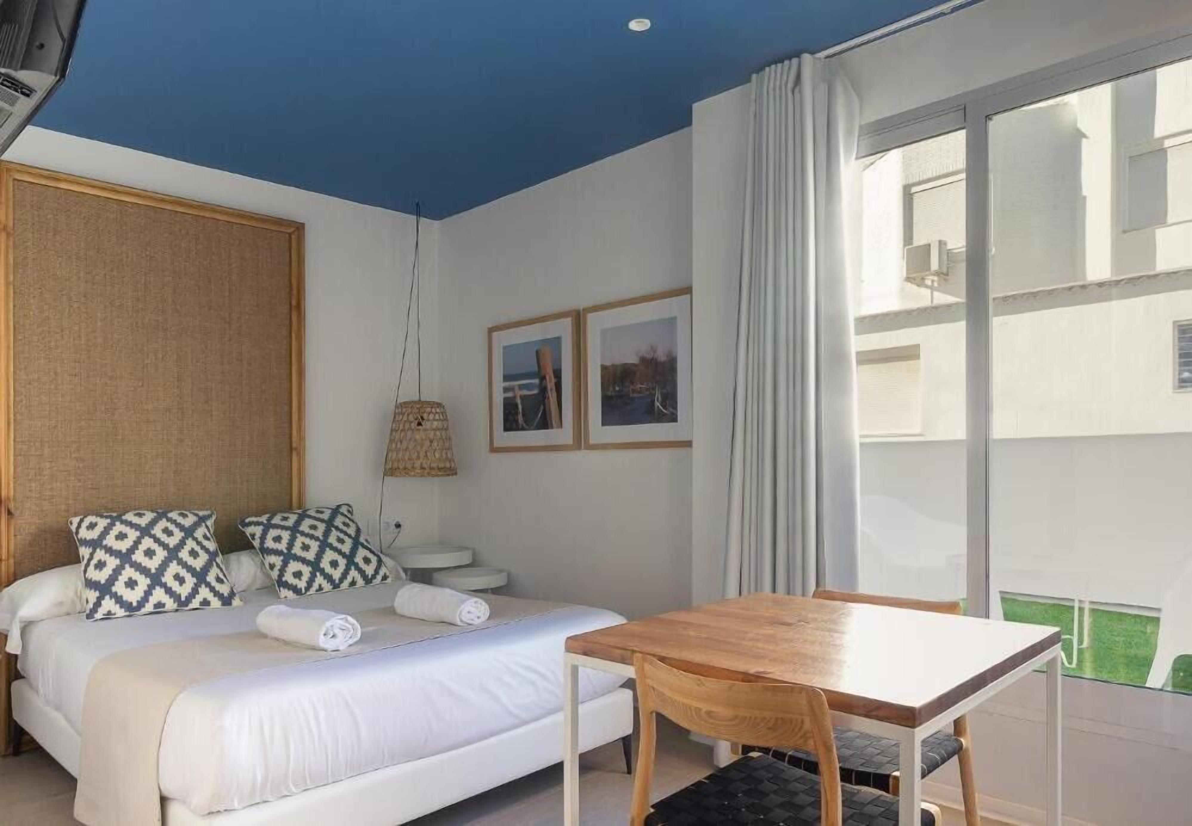 A bedroom with a bed, bedside tables, a wooden table, and a large window.