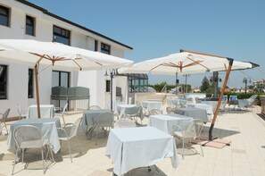 An outdoor patio with tables and chairs, covered by white umbrellas, and a building in the background. - La Sorgente (Capaccio-Paestum)
