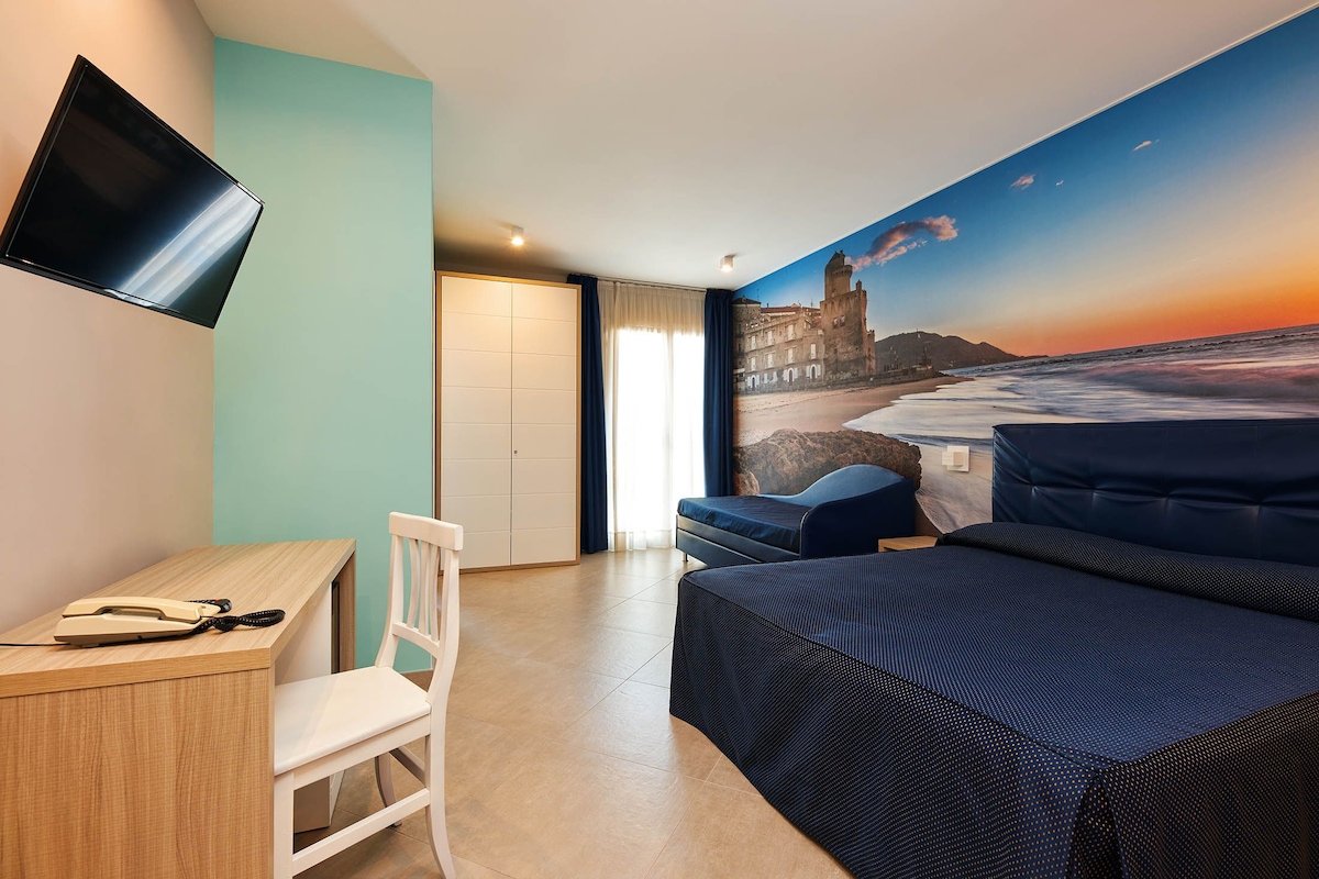 A hotel room with a bed, a desk, a chair, a TV mounted on the wall, and a mural of a beach scene.