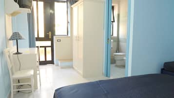 Una camera da letto con un letto, una scrivania con lampada, un armadio e un bagno parzialmente visibile attraverso una porta aperta.
