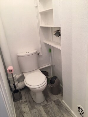 A small, compact bathroom with a toilet, a broom, and a trash bin.