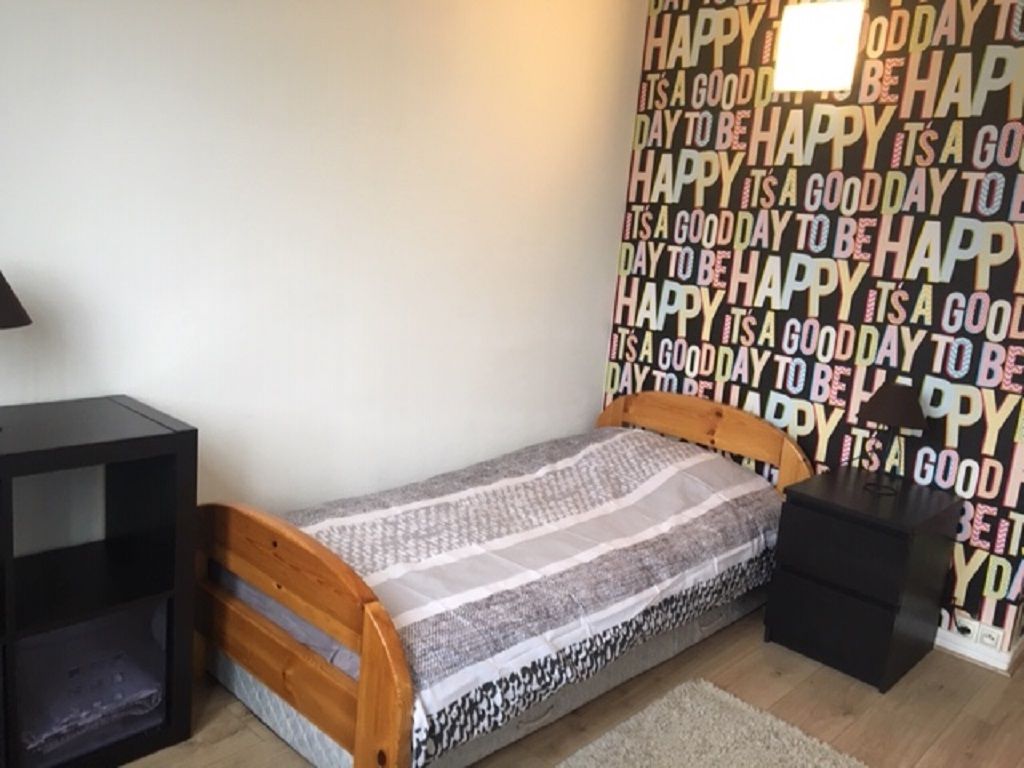 Ein kleines Zimmer mit einem Einzelbett, einem Holz-Kopfstück, einem Nachttisch und einer Wand mit einem sich wiederholenden Muster des Wortes 'HAPPY'.