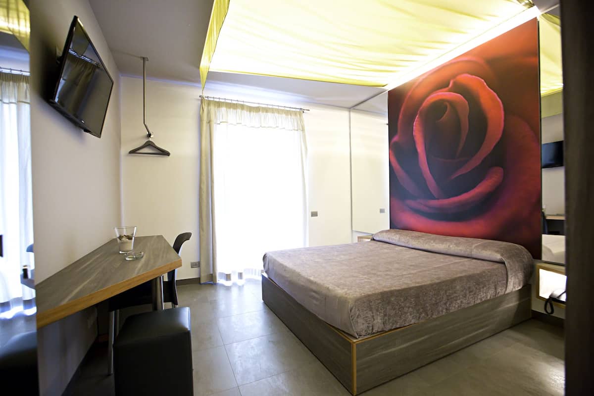 A bedroom with a bed, a desk, a chair, a TV mounted on the wall, and a large rose mural.