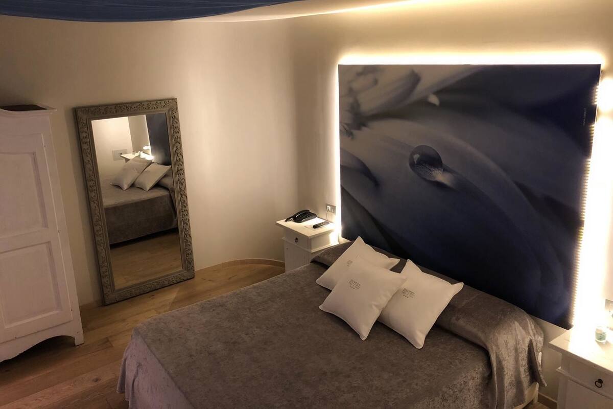 A bedroom with a bed, a nightstand, a mirror, and a wall with a textured artwork.