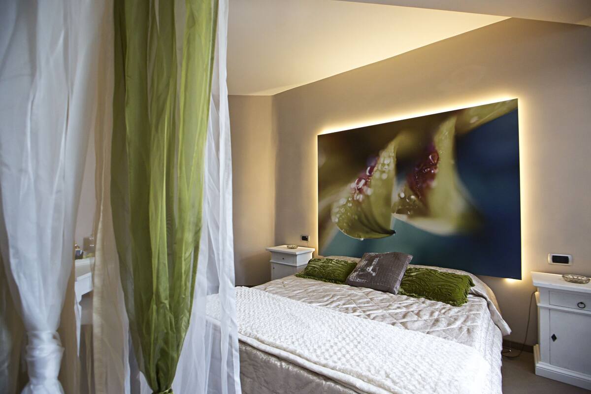 A bedroom with a bed, a nightstand, a wall-mounted lamp, and a large abstract painting.