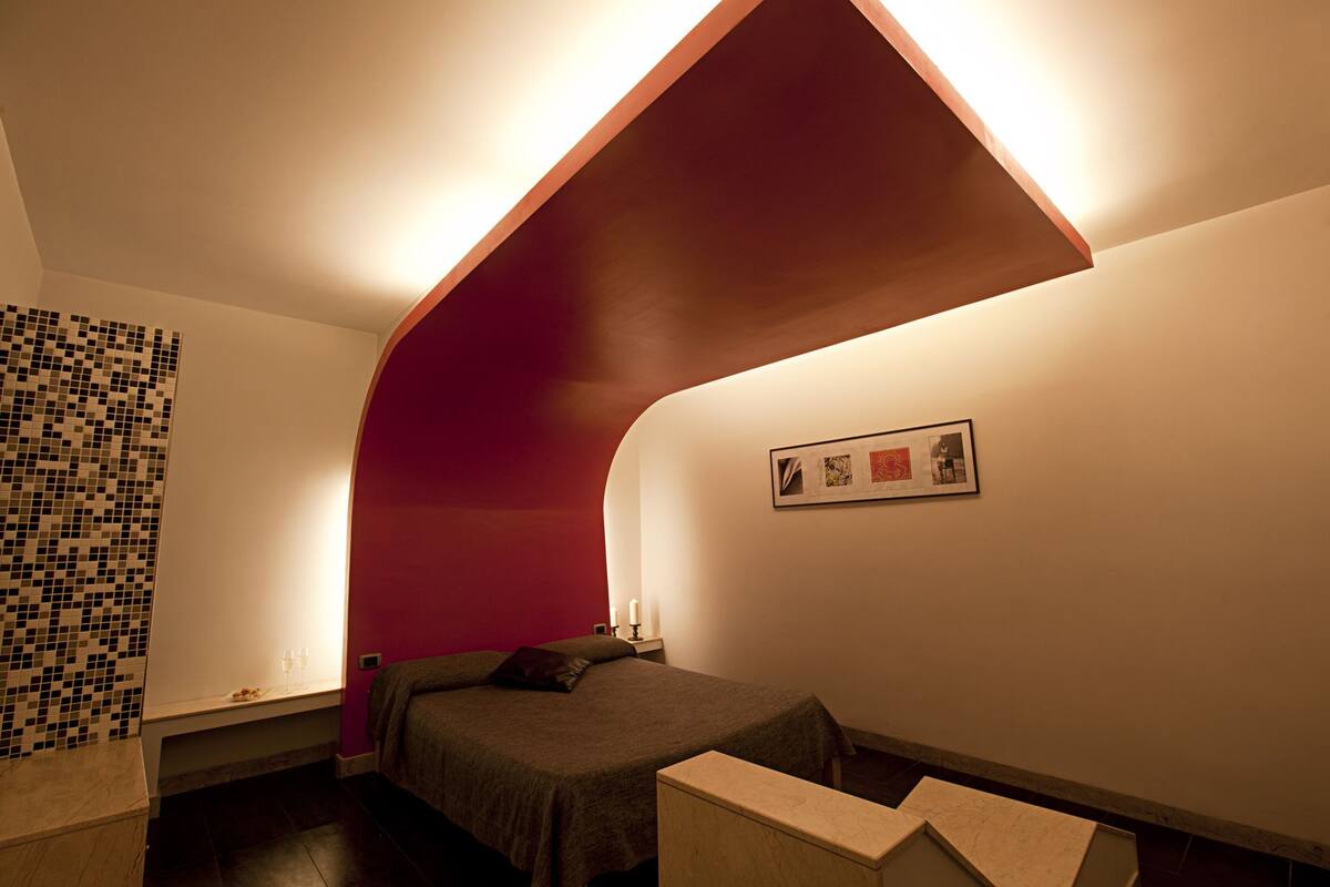 A bedroom with a bed, a nightstand, a wall-mounted picture, and a unique ceiling design.