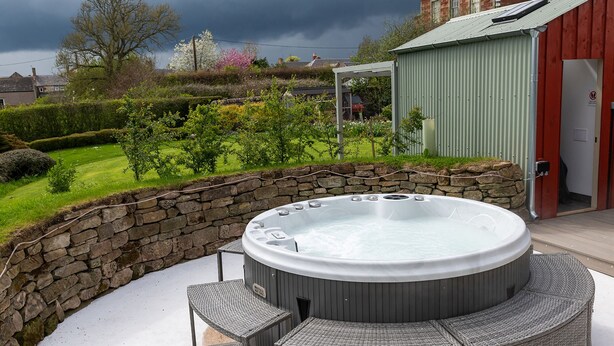 A hot tub with a stone wall and wicker furniture, surrounded by a garden and a red building in the background. - A beautiful and cosy dog friendly cottage with flexible sleeping arrangements (Greenlaw)