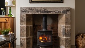 A stone fireplace with a modern wood-burning stove, a wooden table with a vase and books, and a wicker basket on the floor. - A beautiful and cosy dog friendly cottage with flexible sleeping arrangements (Greenlaw)