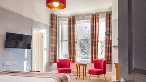 Habitación de hotel con cama, dos sillas rojas, una mesita, televisor y ventana con cortinas.