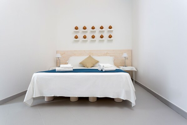A hotel room with a neatly made bed, white bedding, a blue accent throw, a wooden headboard, a beige pillow, and a wall-mounted shelf with decorative items. - Hotel L'Uomo e il Mare (Gallipoli)