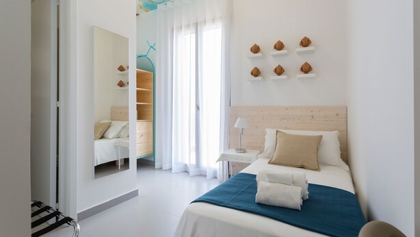 Habitación de hotel moderna con cama, escritorio y silla. Se observa una ventana con cortinas, un espejo y una estantería con adornos.