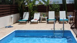 A swimming pool with lounge chairs and a table, surrounded by a wooden fence and plants. - Vasilisa villa BW16 (Paralimni)