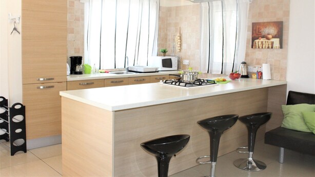 A modern kitchen with a central island, bar stools, and a dining area with a sofa. - Vasilisa villa BW16 (Paralimni)