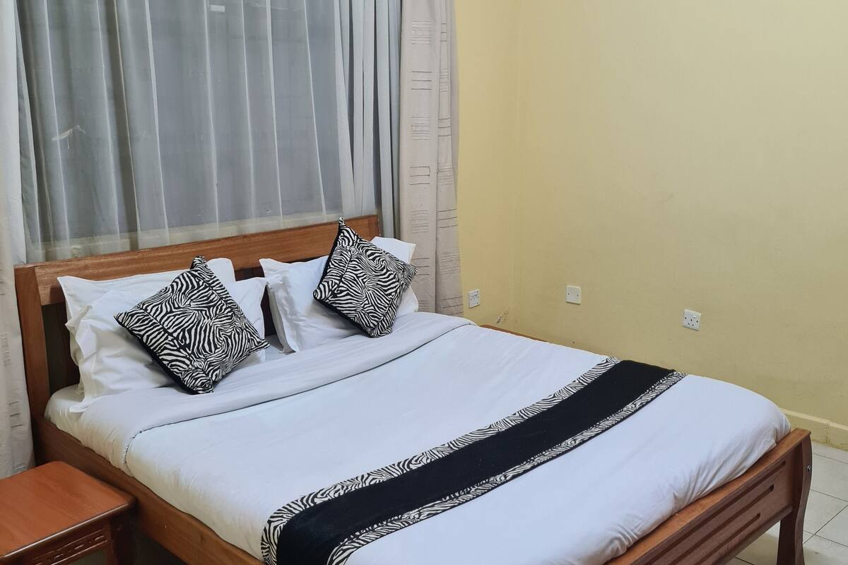 Ein ordentlich bezogenes Bett mit weißer Bettwäsche und Kissen mit Zeprazebra-Muster, ein hölzernes Kopfteil und ein Nachttisch.