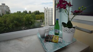 A window view of a cityscape with buildings and trees, a table with a bottle, a cup, and some snacks. - Elegant Apartment Arkadia (Warsaw)