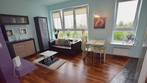 A modern living room with a sofa, dining table, and chairs. There are large windows with a view of buildings outside. - Elegant Apartment Arkadia (Warsaw)