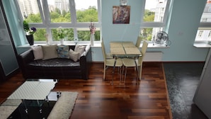 A living room with a sofa, a dining table with chairs, a fan, and a window with a view of buildings and trees. - Elegant Apartment Arkadia (Warsaw)