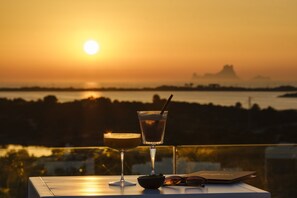 Una mesa con una bebida, un bol y un libro frente a una vista del atardecer.