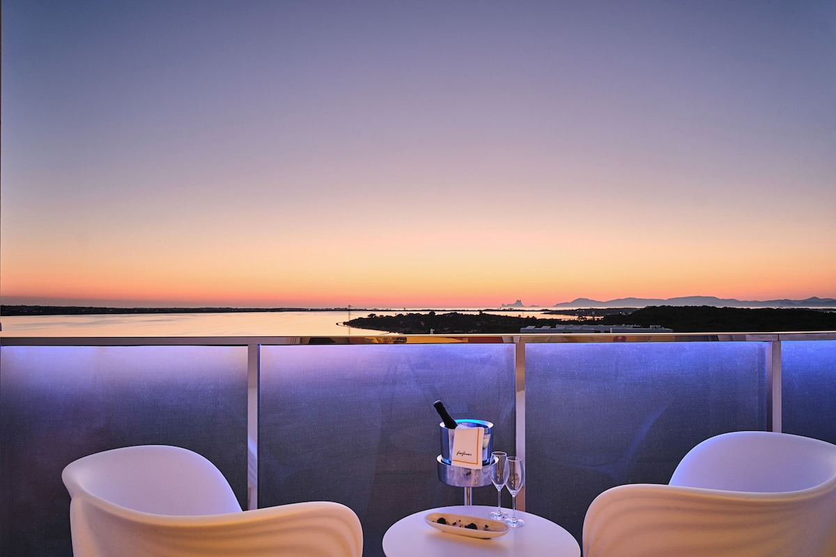 A balcony with two white chairs, a table with a bottle of champagne, and a glass of wine.