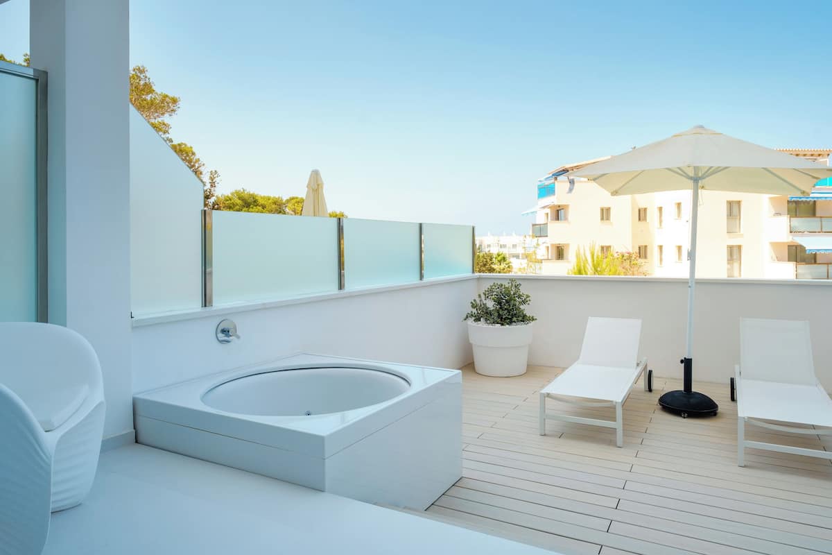 A modern outdoor bathroom with a bathtub, two white lounge chairs, a potted plant, and a large umbrella.